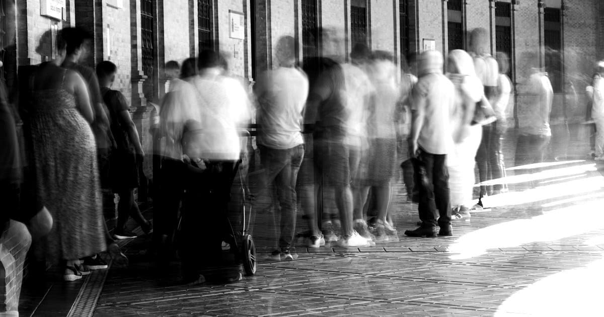 Cola de personas esperando en fila frente a un edificio, difuminadas por el paso del tiempo.